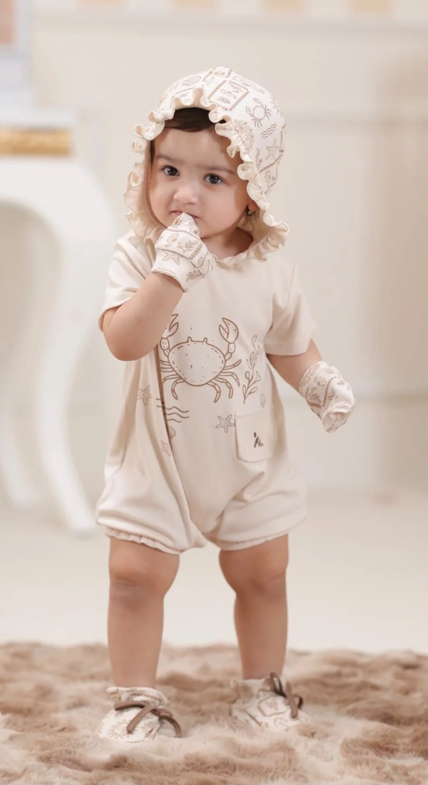 A young child wearing a cream illustrated Pythal romper and matching bonnet.